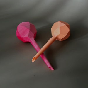 Op de afbeelding: Twee plastic gietkannen met geometrische vormen. Eén is roze en de andere is oranje.