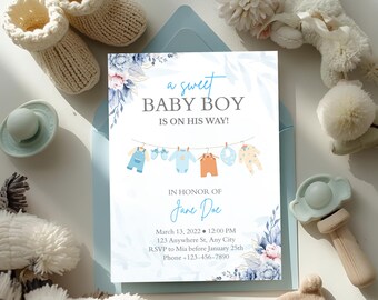 May include: Baby shower invitation with the text "a sweet BABY BOY IS ON HIS WAY!" in blue script. The invitation features baby clothes hanging on a clothesline, floral accents, and event details. Soft blue and cream-colored baby items surround the invitation.