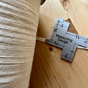 May include: A spool of beige woven ribbon next to a metal measuring gauge. The gauge reads "Measuring Gauge" and indicates measurements in inches. The ribbon appears to be approximately 2.5 cm wide.