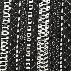 May include: Close-up of a black lace fabric with a repeating pattern. The design features vertical stripes with geometric and floral elements. The lace is detailed with openwork and varying textures, creating a visually rich surface.