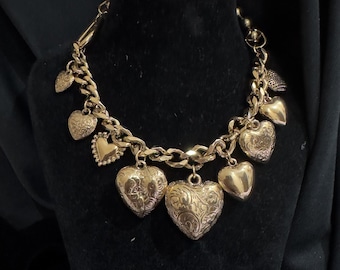 May include: A gold-coloured charm bracelet with a chunky chain and various heart-shaped charms. Some hearts are solid, others have detailed engraved patterns. The bracelet is displayed on a black velvet surface.