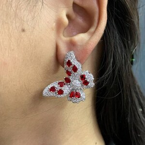 Può includere: Un orecchino a farfalla argentato con pietre preziose rosse e trasparenti. Il design a farfalla presenta pietre preziose rosse al centro delle ali e pietre preziose trasparenti che delineano le ali. L'orecchino è a lobo.