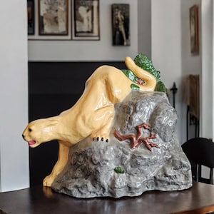 May include: A ceramic sculpture of a yellow cougar crouching on a gray rock with green foliage. The cougar is positioned as if it is about to pounce.