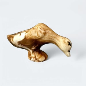 Op de afbeelding: Een keramiek eendenbeeld met bruine en witte marmering. De eend zit in een hurkende positie met zijn kop naar beneden, alsof hij drinkt.