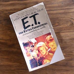 May include: A paperback book cover with a yellow background and the title "E.T. The Extra-Terrestrial" in black text. The cover features a photo of three children, two boys and a girl, looking at the camera. The book is based on the Steven Spielberg film.