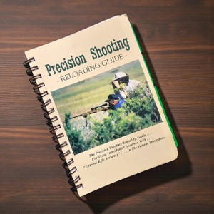 May include: A spiral-bound book titled "Precision Shooting - Reloading Guide" with a green spine. The cover features a photograph of two people aiming a rifle. The text below the image reads "For Those Individuals Concerned With...Extreme Rifle Accuracy..."