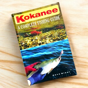 以下が含まれることがあります： 黄色と赤色のタイトルが付いた「Kokanee」というタイトルの完全な釣りガイド。表紙にはルアーで釣られた魚の写真が掲載されています。本は明るい木製の表面にあります。