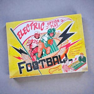 以下が含まれることがあります： 黄色い背景のヴィンテージなElectric Option Play Footballボードゲームの箱。カバーには、アクション中の2人のフットボール選手、スタジアムのグラフィック、そしてElectric Option Play Footballと書かれた太字のテキストが描かれています。