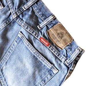May include: A close-up of the back pocket of a pair of blue denim jeans. The pocket has a red "Wrangler" label and a brown leather patch with the Wrangler logo and "Premium Quality" text.