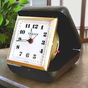May include: A vintage black travel clock with a white face, gold trim, and black hands. The clock face displays the word "EQUITY" and the numbers 1 through 12. The clock is in a black case.