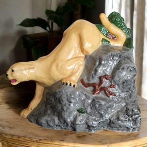 May include: A ceramic figurine of a light brown cougar crouching on a gray rock with brown branches. The cougar's tail is curled around the rock.