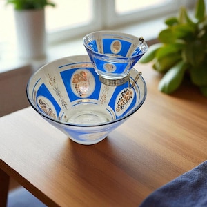 May include: A vintage glass serving bowl set with a smaller bowl resting on top. The bowls feature a blue and white design with gold grape and wheat motifs. The set is displayed on a wooden table.