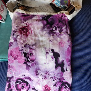 May include: A tote bag with a floral pattern in shades of purple and pink. The bag has a white canvas top and a vibrant floral print on the main body, showcasing roses and other flowers.