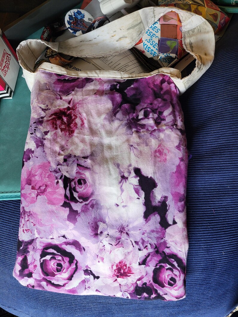 May include: A tote bag with a floral pattern in shades of purple and pink. The bag has a white canvas top and a vibrant floral print on the main body, showcasing roses and other flowers.