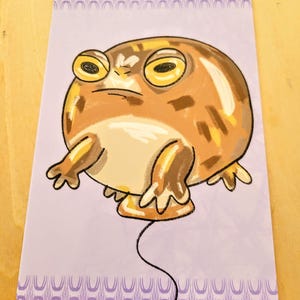 Puede incluir: Ilustración de una rana con expresión de enfado, en tonos marrones y beige, con grandes ojos amarillos. La rana está sobre un fondo lavanda con un borde decorativo. Una línea negra sale del cuerpo de la rana.
