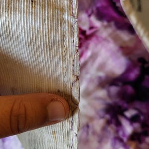 May include: Close-up of fabric edges. The fabric on the left is a light beige with vertical lines and some dark spots. The edge is sewn with purple thread. The background is a purple and white floral pattern.