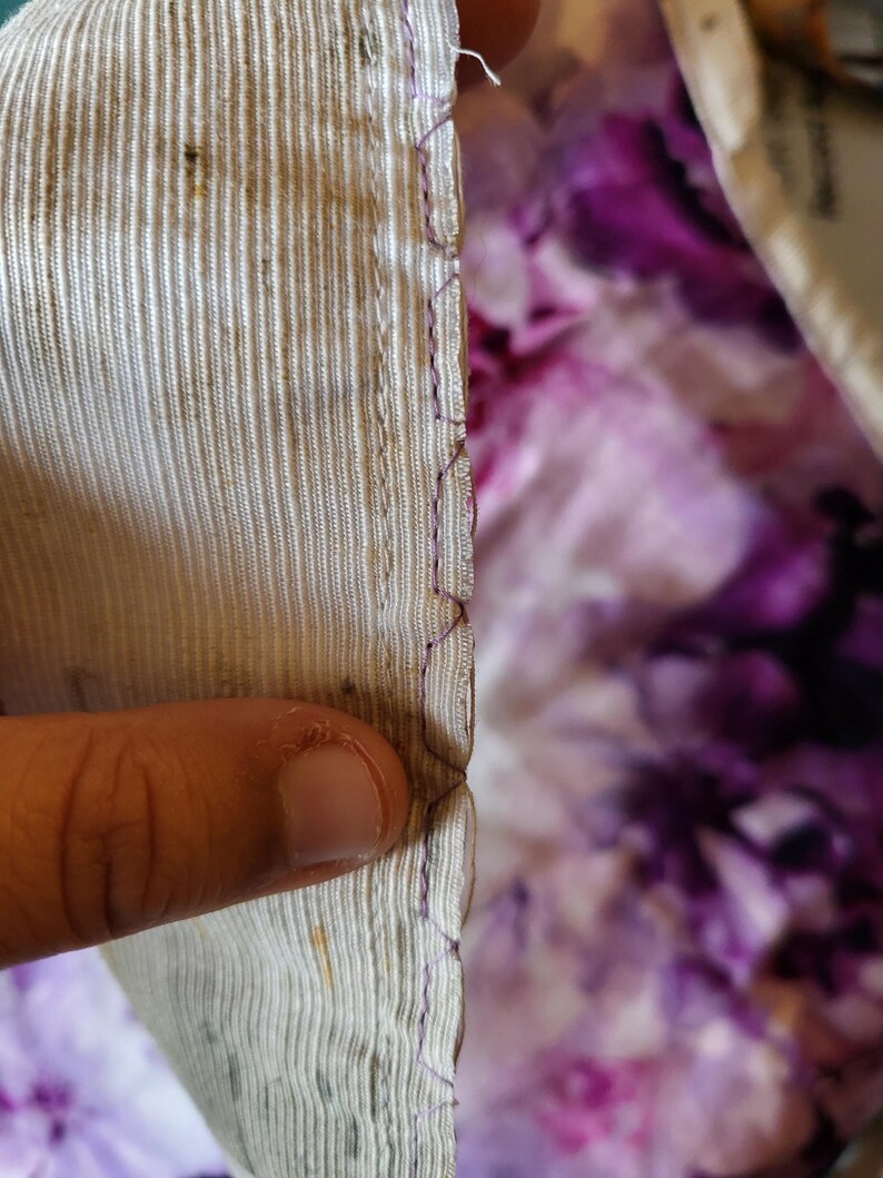May include: Close-up of fabric edges. The fabric on the left is a light beige with vertical lines and some dark spots. The edge is sewn with purple thread. The background is a purple and white floral pattern.