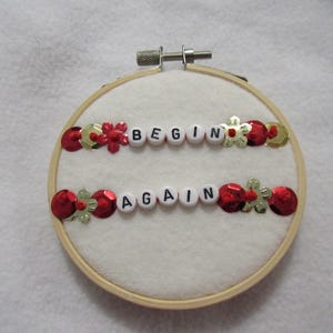 May include: A round embroidery hoop with the words "BEGIN AGAIN" spelled out in white letter beads. The words are above and below each other. Red sequins and gold flower-shaped sequins decorate the top and bottom of the words.