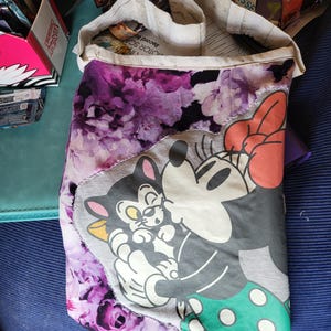 May include: A handmade tote bag with a floral purple pattern and a grey panel featuring cartoon characters. The bag's design includes a Minnie Mouse graphic with a red bow and a green polka-dot skirt. The bag is open, revealing its interior.