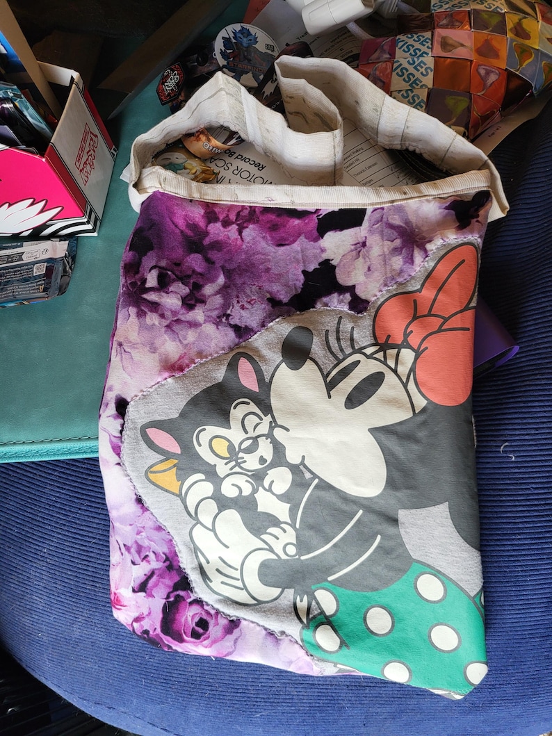 May include: A handmade tote bag with a floral purple pattern and a grey panel featuring cartoon characters. The bag's design includes a Minnie Mouse graphic with a red bow and a green polka-dot skirt. The bag is open, revealing its interior.