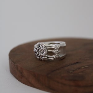 May include: A silver ring crafted from a spoon, featuring a floral design on the face. The ring is displayed on a dark wooden surface, showcasing its intricate details and vintage aesthetic. The ring's band is smooth and polished.