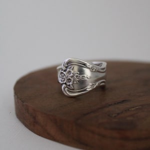 May include: A silver spoon ring with a floral design. The ring is made from a repurposed spoon and features intricate details. The ring is displayed on a wooden surface.