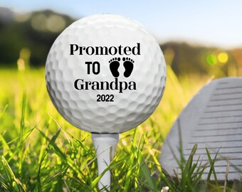 May include: White golf ball with black text that reads "Promoted to Grandpa 2022" with a pair of black footprints. The golf ball is sitting on a tee in green grass.