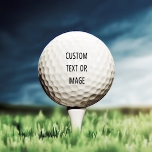 Pode incluir: Uma bola de golfe branca com o texto "CUSTOM TEXT OR IMAGE" nela, sentada em um tee verde em um campo gramado.