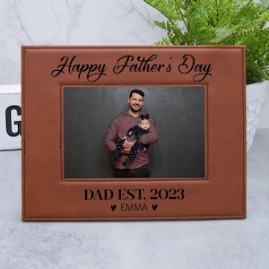 May include: Brown leather photo frame with a picture of a father and child. The frame has the text "Happy Father's Day" at the top and "DAD EST. 2023 ♥ EMMA ♥" at the bottom.