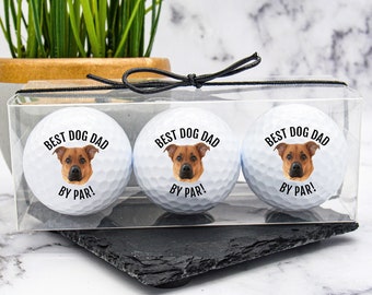 May include: Three white golf balls with a black and white image of a dog and the text "Best Dog Dad By Par!" printed on each ball. The golf balls are in a clear plastic box with a black ribbon.