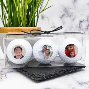 May include: Three white golf balls with personalized photos in a clear plastic box with a black ribbon.