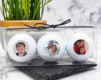 Può includere: Tre palline da golf bianche con foto personalizzate in una scatola di plastica trasparente con un nastro nero.