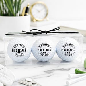 May include: A clear box containing three white golf balls. Each ball is printed with the text "KEVIN JAMES RING BEARER 03-07-2021" in black. The box is tied with a black ribbon. Golf accessories are in the foreground.