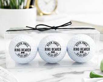 May include: A clear box containing three white golf balls. Each ball is printed with the text "KEVIN JAMES RING BEARER 03-07-2021" in black. The box is tied with a black ribbon. Golf accessories are in the foreground.