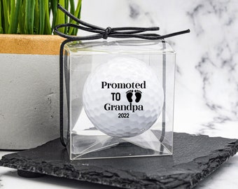 May include: A white golf ball in a clear box with a black ribbon. The golf ball has the text "Promoted to Grandpa 2022" and a pair of baby footprints.