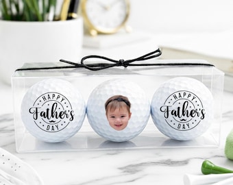 May include: A set of three white golf balls in a clear box, tied with a black ribbon. Two balls have a "Happy Father's Day" design, and the center ball features a child's photo. Golf accessories are in the foreground.
