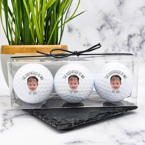 May include: Three white golf balls in a clear plastic case. Each ball features a child's face and the text "THE GODFATHER OF MIA EST. 2026". The case is tied with a black ribbon. The golf balls are in a clear plastic case.