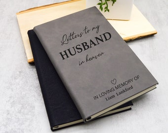 May include: A gray journal with the text "Letters to my HUSBAND in heaven" and "IN LOVING MEMORY OF Liam Lankford" is stacked on top of a black journal. A small white heart is below the text. A small succulent plant in a white square pot is in the background.