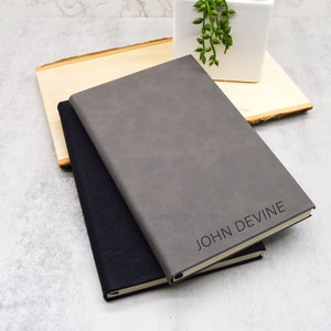 May include: Two leather-bound journals, one gray and one black, with the name "JOHN DEVINE" embossed on the gray journal. The journals are stacked on top of each other and are sitting on a wooden surface.