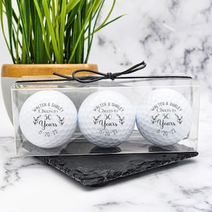 May include: Three white golf balls in a clear plastic box with a black ribbon. Each ball is personalized with the text "Walter & Shirley Cheers to 50 Years 8-20-22".