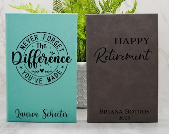 May include: Two personalized leather journals. The teal journal has the text "Never Forget The Difference You've Made" and the name "Lauren Schecter". The gray journal has the text "Happy Retirement" and the name "Briana Botros 2023".