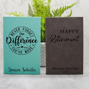 May include: Two personalised leather journals. The teal journal has the text "Never Forget The Difference You've Made" and the name "Lauren Schecter". The grey journal has the text "Happy Retirement" and the name "Briana Botros 2023".
