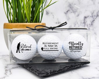 May include: Three white golf balls in a clear plastic box with a black ribbon. The golf balls are decorated with black text that reads "Retired and loving it", "Retirement is now in full swing", and "Officially Retired let the fun begin".
