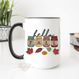May include: White ceramic mug with a black handle. The mug has a design of four mason jars with the word "FALL" spelled out on the jars. The jars are decorated with fall colors and have twine tied around the lids. The text "hello" is written above the jars. There are also fall leaves scattered around the jars.