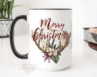 May include: White ceramic mug with a black handle and rim. The mug features the words "Merry Christmas" in a burgundy script, along with antler and floral illustrations. Gold glitter accents the design.