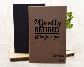 May include: A brown leather-bound journal with the text "Officially Retired Let the fun begin" and the name "Cory Moore" and the year "2023" engraved on the front. The journal is sitting on a wooden surface.