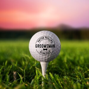 May include: A white golf ball with a black circle sticker that says "PAYTON PHILLIPS GROOMSMAN 07.08.2023" with a bow tie icon.