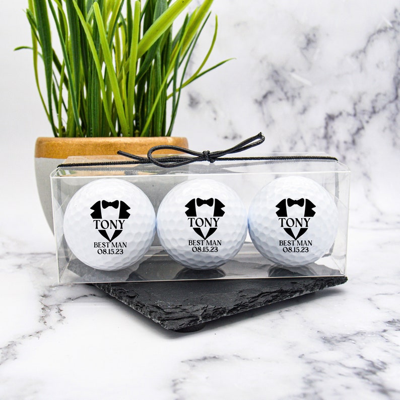 K&ouml;nnte beinhalten: Drei wei&szlig;e Golfb&auml;lle in einer durchsichtigen Plastikbox mit einem schwarzen Band. Jeder Ball hat eine schwarze Smokinggrafik mit dem Text "TONY BEST MAN 08.15.23"