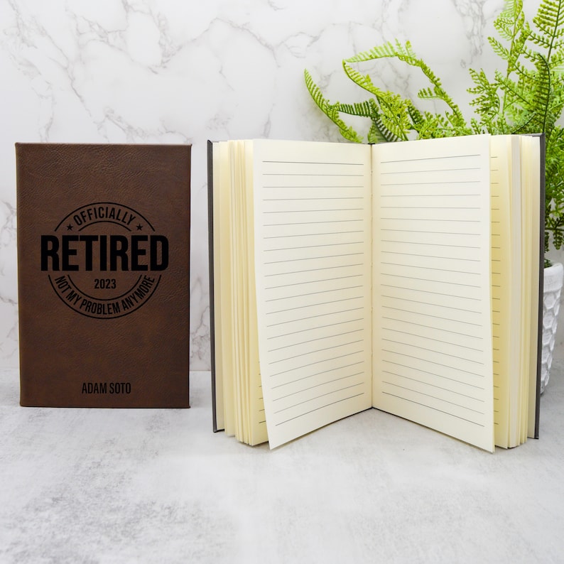 May include: A brown leather-bound journal with the text "Officially Retired 2023 Not My Problem Anymore" and the name "Adam Soto" printed on the front cover. The journal is open to reveal lined pages.