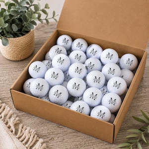 May include: A cardboard box filled with white golf balls, each featuring a black printed monogram with the letter M and the name MICHAEL IVI. The golf balls are arranged in a grid pattern inside the open box, showcasing the personalized design.